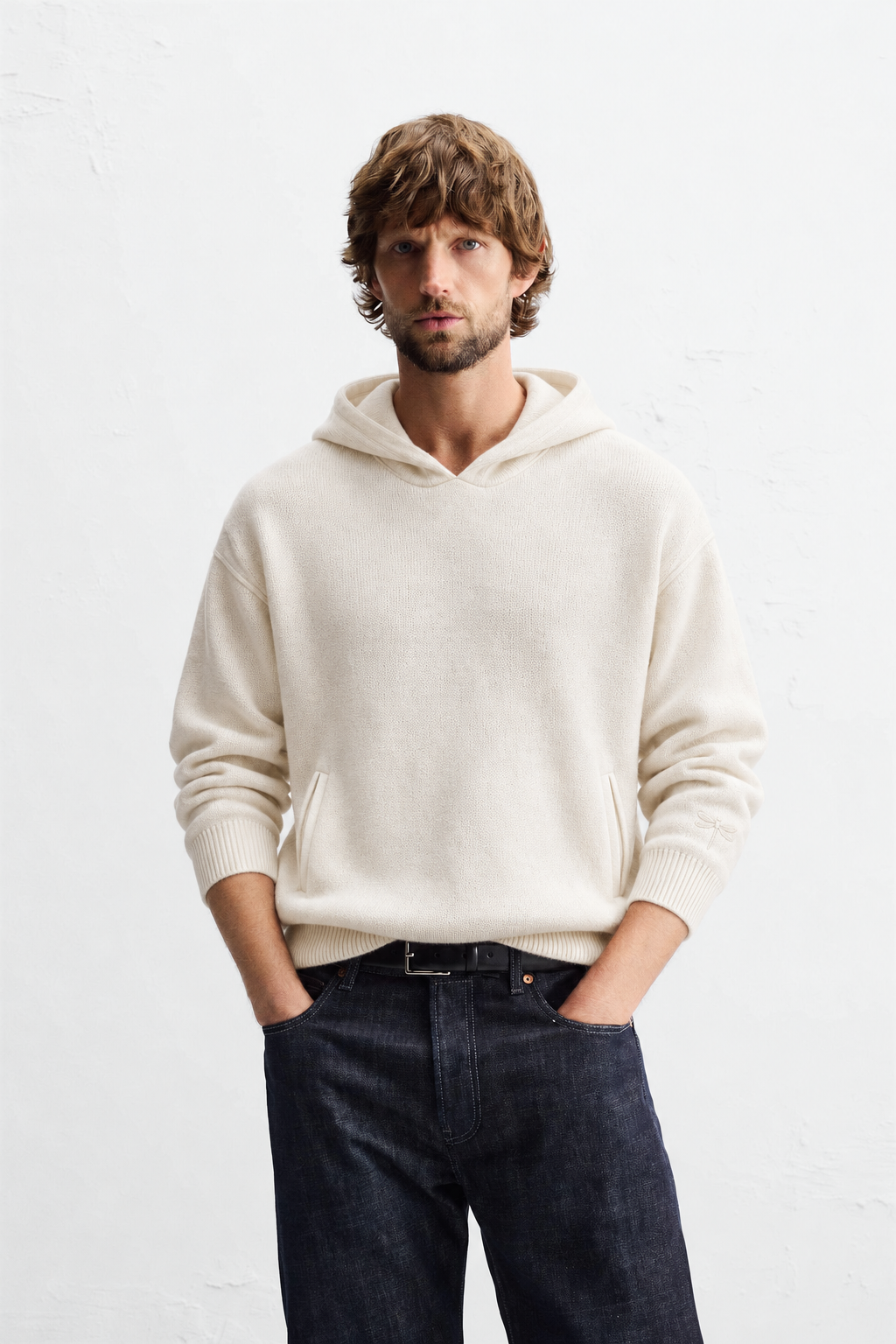 A man wearing an ivory knit hoodie and dark jeans
